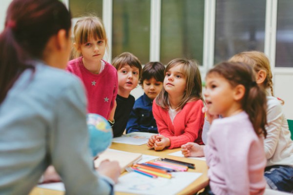 Preschool teacher teaching her children about geography. Using globe and asking the questions. Children  answering the questions. Children sitting by the table. Selective focus to girl in the middle. Models in this shot are part of real kindergarten group and their teacher.