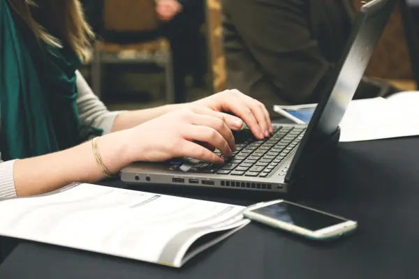 Person typing on a laptop on a desk, next to a mobile phone and some documents.