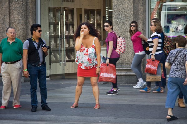 A scene from 'Poses' featuring a woman standing frozen on a sidewalk, emulating a model's pose. She draws looks from passing pedestrians. (Photo: Wikimedia Commons)