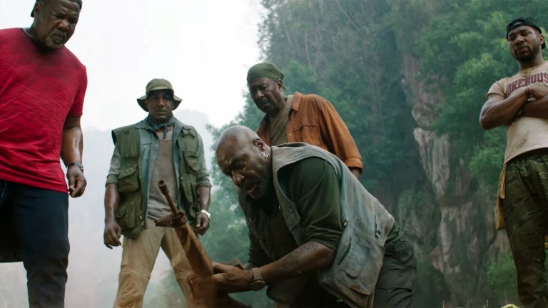 One of Da 5 Bloods kneels down inspecting a gun in a Vietnamese jungle. The other four Bloods stand over him also looking at the gun.