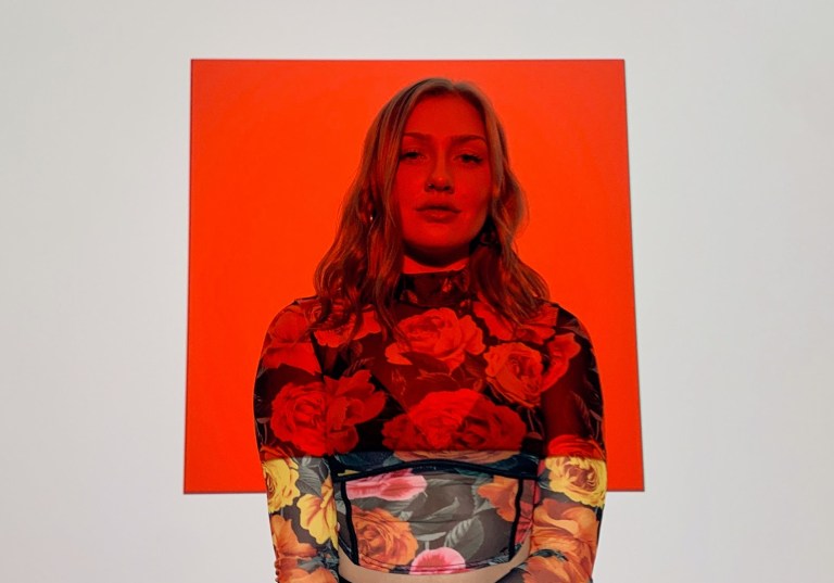 Olivia Lunny stands against an off-white background with a red square projected over her face and upper torso. She wears a sheer floral long-sleeve.