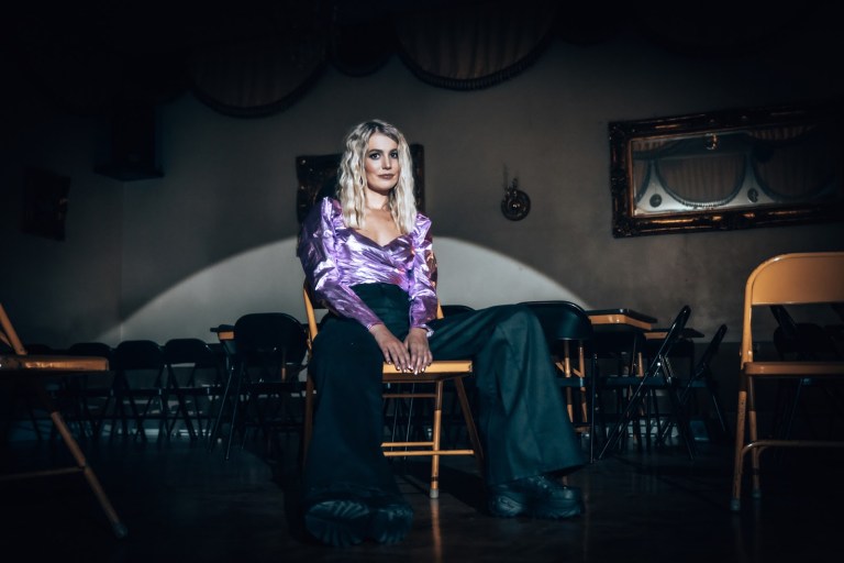 Cali Rodi sits on a metal folding chair. In the dimly lit background are several more folding chairs and tables.