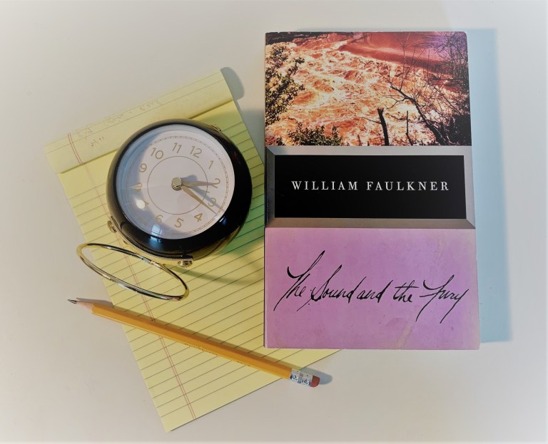 A desk arrangement of a copy of William Faulkner's "The Sound and the Fury," a clock, a pencil, and a notepad