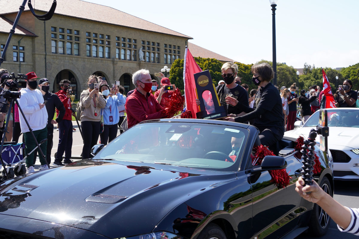 Tara VanDerveer sits in a convertable and hands Marc Tessier-Lavigne the NCAA women's basketball trophy as students watch and take pictures.