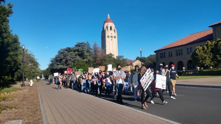 Students march down Palm Drive with Hoover Tower in the background. All are wearing masks and some are wearing signs condemning the Israeli occupation of Palestine.