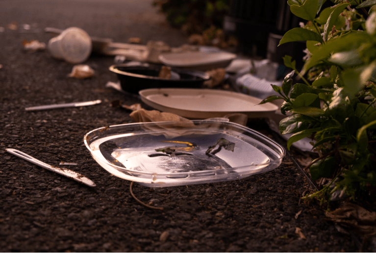 Close up on plastic container lid, knives, cups and bottles in a pile on concrete near a black trashcan and green bush