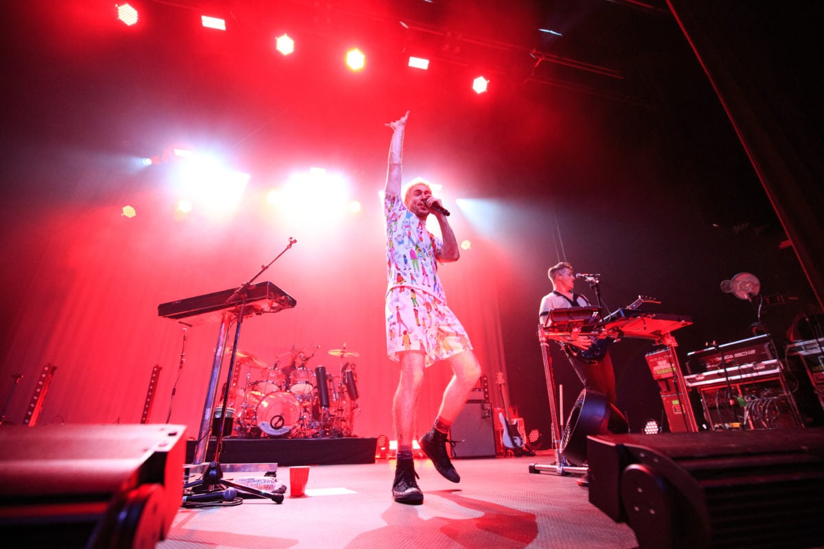 Wide image of Nicholas Petricca singing on a red-lit stage with a drum set behind him and instrumentalist to the right of the frame.