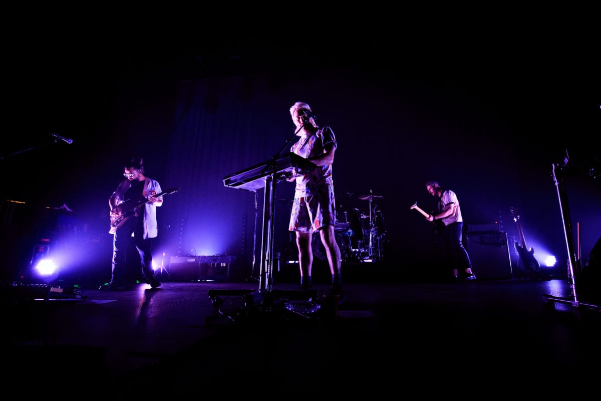 Medium image of all of the band members of Walk the Moon on a dark stage.