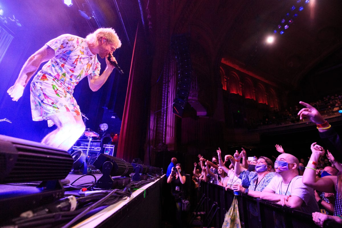 Wide image of Nicholas Petricca singing, bent over, on a blue-lit stage towards an illuminated crowd.