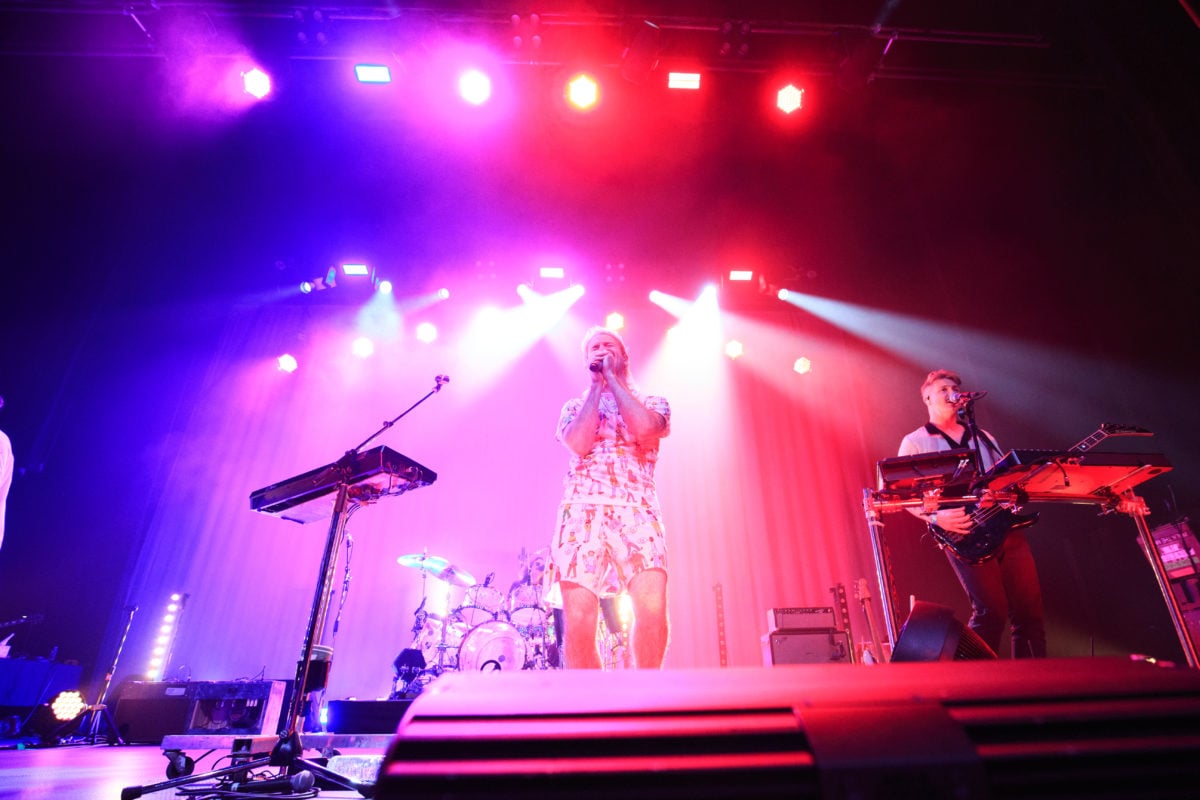 Wide image of Nicholas Petricca singing on a blue, white and red-lit stage with an instrumentalist to the right of the frame.