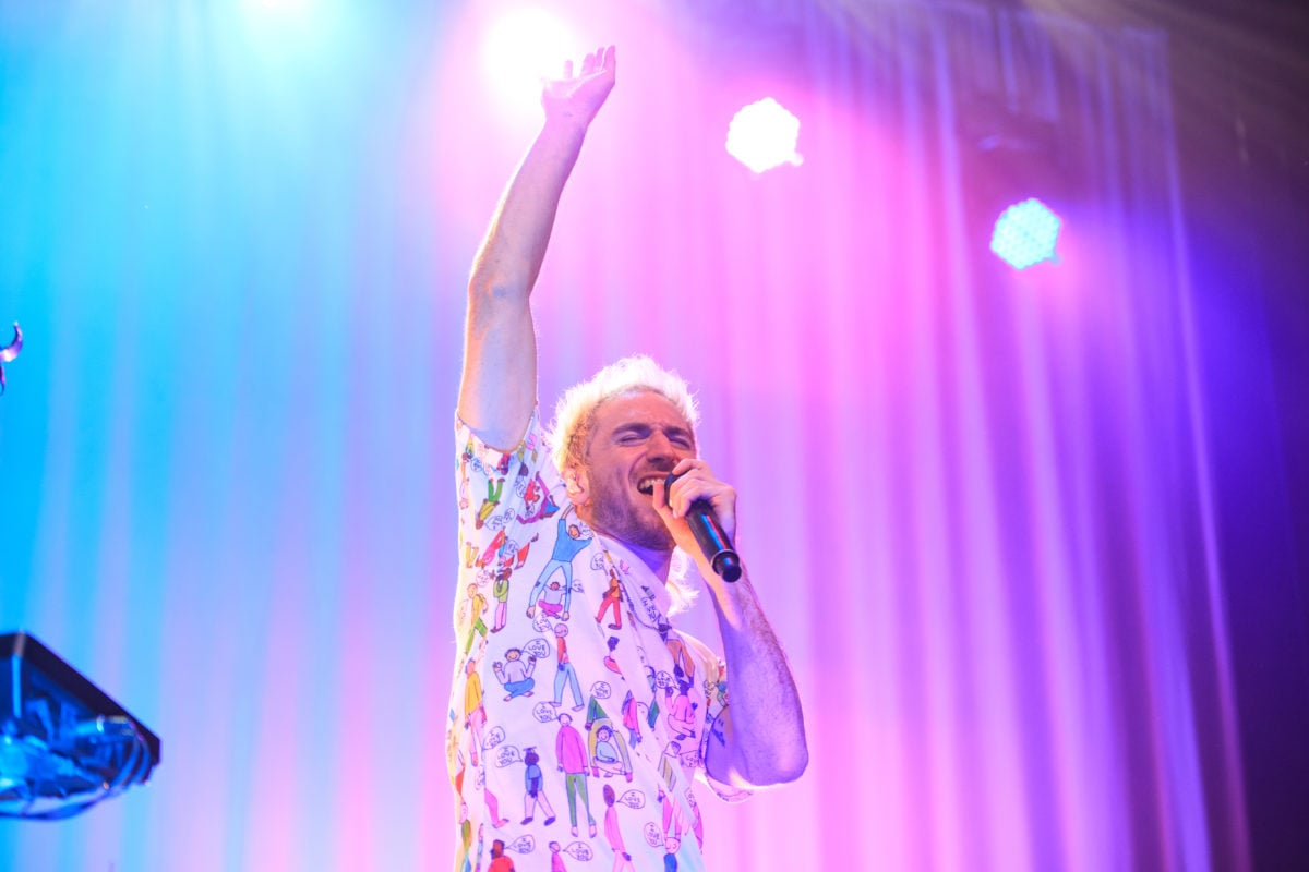 Medium image of Nicholas Petricca singing with his hand stretched upwards on a blue, purple and pink-lit stage.