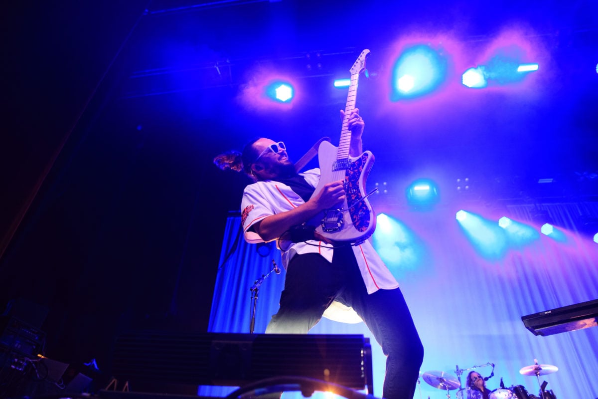 Wide image of guitarist Eli Maiman playing guitar on a blue-lit stage. A drum set can be seen in the lower right corner of the frame.