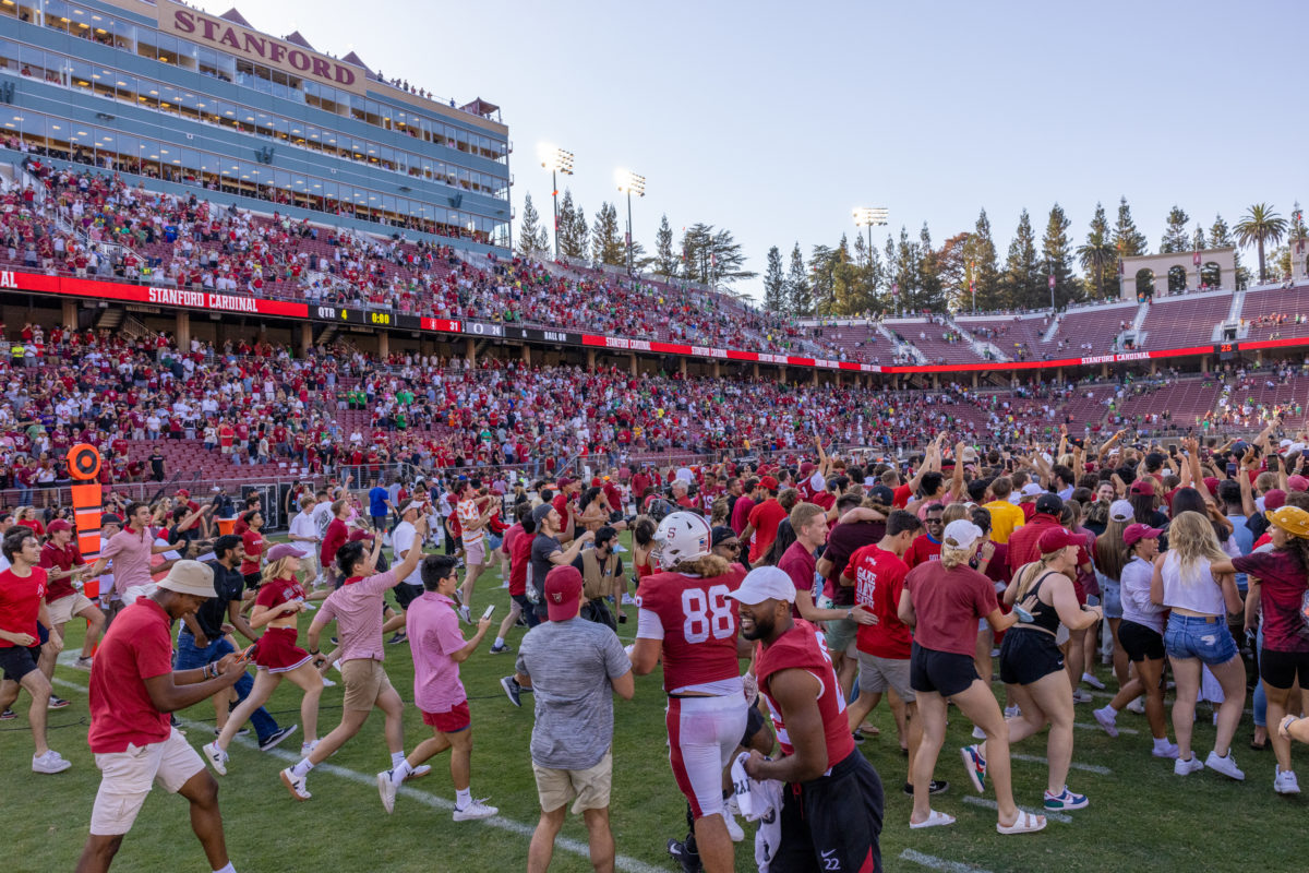 A large crow of Stanford fans, many wearing cardinal, storm the field from the left with their arms in the air. Many still stand in the stadium bleachers.