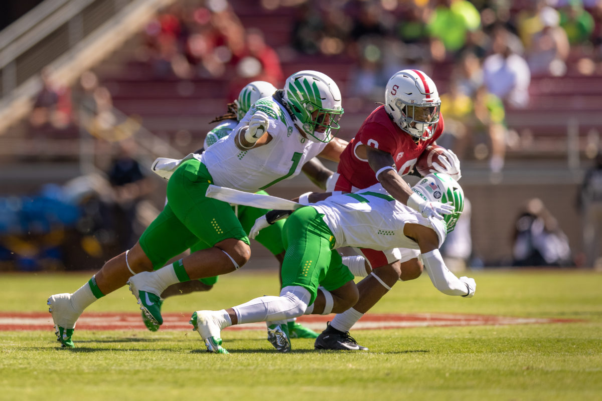 Running back Nathaniel Peat runs to the right with the ball in his left hand. Charging in front of his abdomen, an Oregon football player blocks his path. Right behind him, two more Oregon players, with extended arms, ready to tackle him.