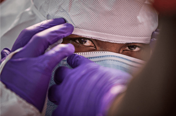 A man wearing a hazmat suit, a mask and purple gloves, staring into the camera.