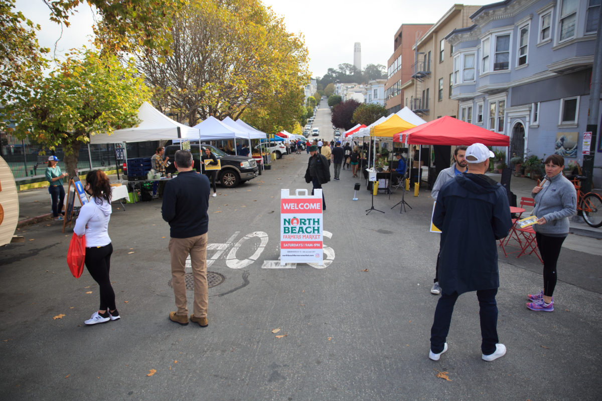 Farmers market sign in middle of farmers market with people, looking down a street