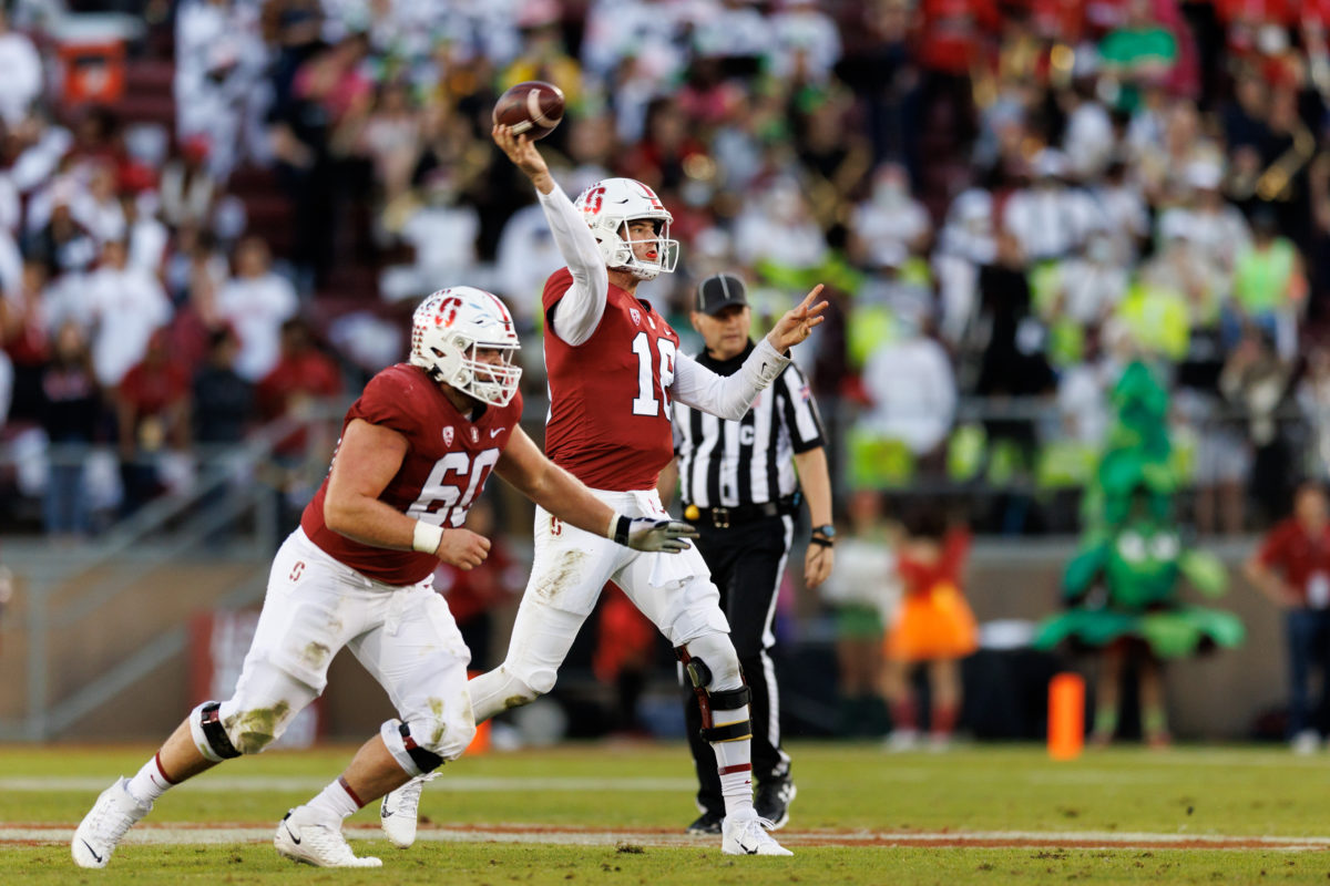 McKee throws a football on the field as another player runs in front of him, referee behind him. 