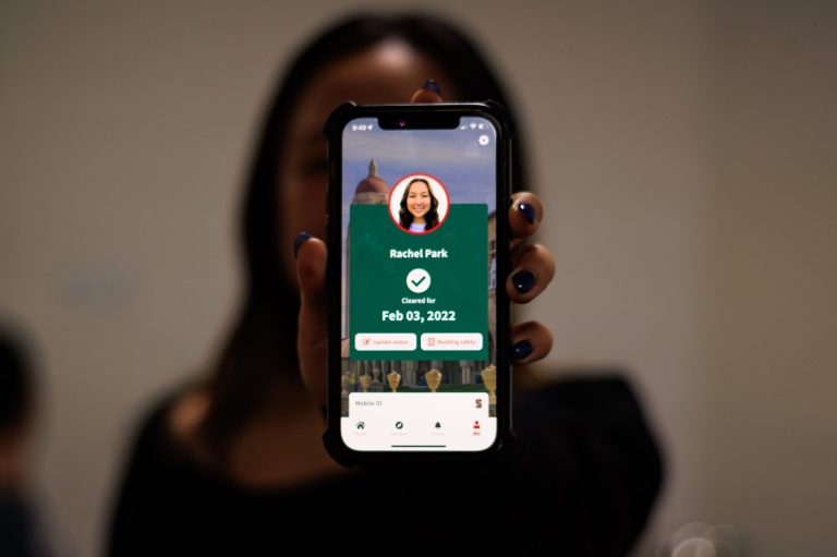 A student stands for a photo, holding out her phone that shows her green onsite access badge