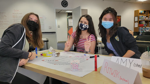 Three students sitting around a table smile at the camera. They are working on a poster for their data science summer camp.