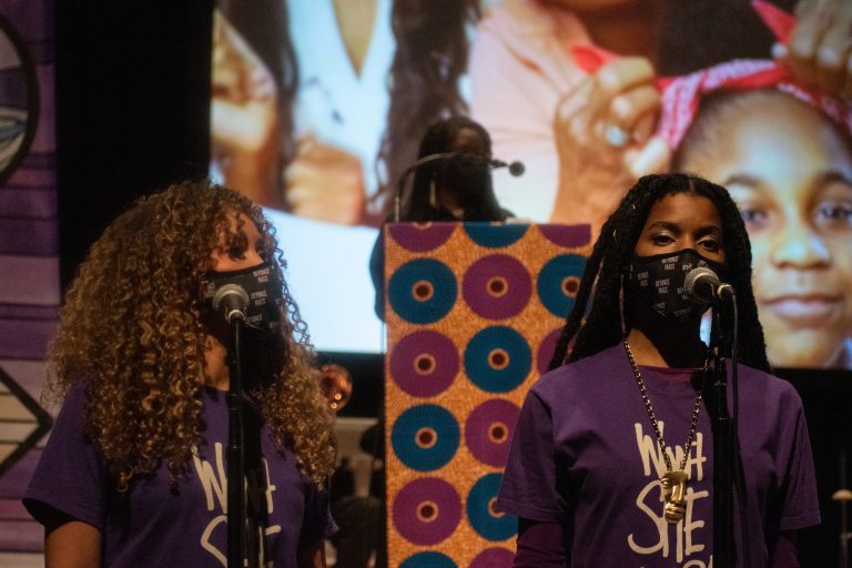 Two women wearing purple shirts that say, "Won't she do it?!" sing in front of microphones.