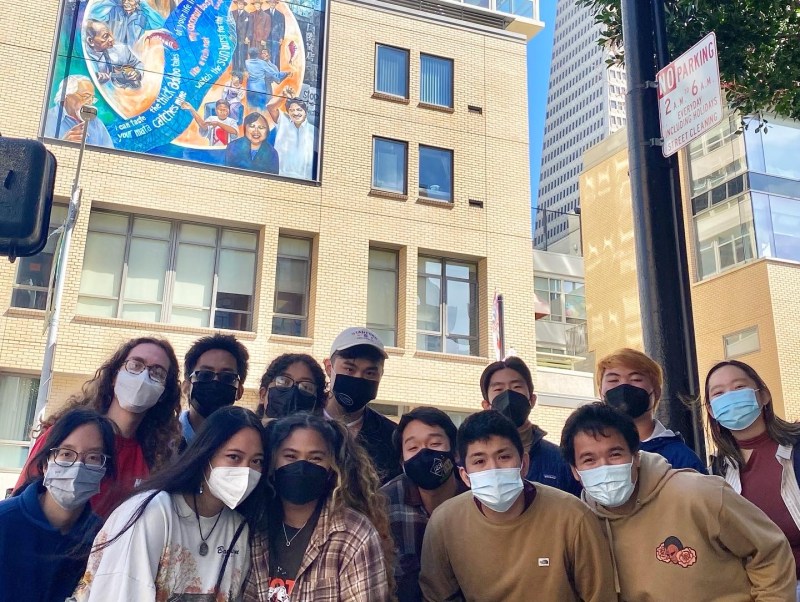 A group of students stand for a photo in front of a building with a mural in downtown San Francisco.