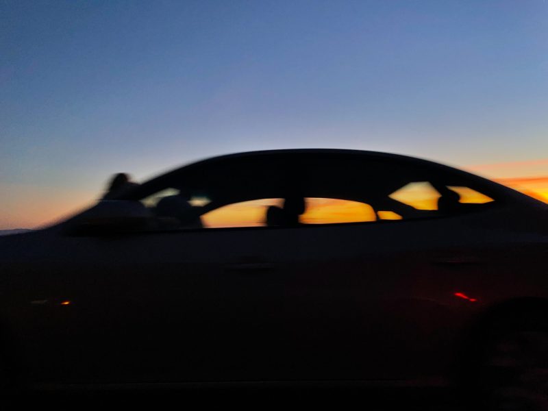 The silhouette of a sedan races by in front of an orange sunset in a deep blue sky.