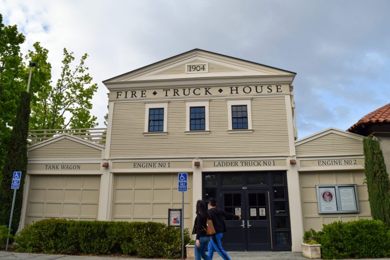 picture of fire truck house, "tank wagon, engine no 1, ladder truck no 1, engine no 2" seen on front of building. two people walking in front.