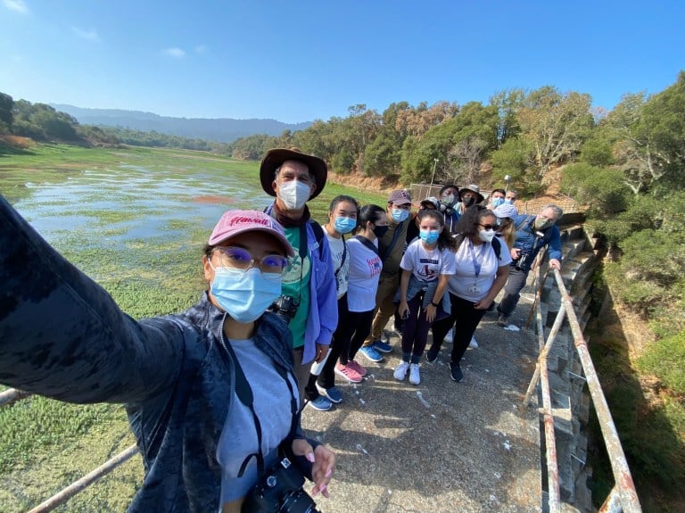 A woman holds up a camera as she takes a photo with a group of students on a bridge.