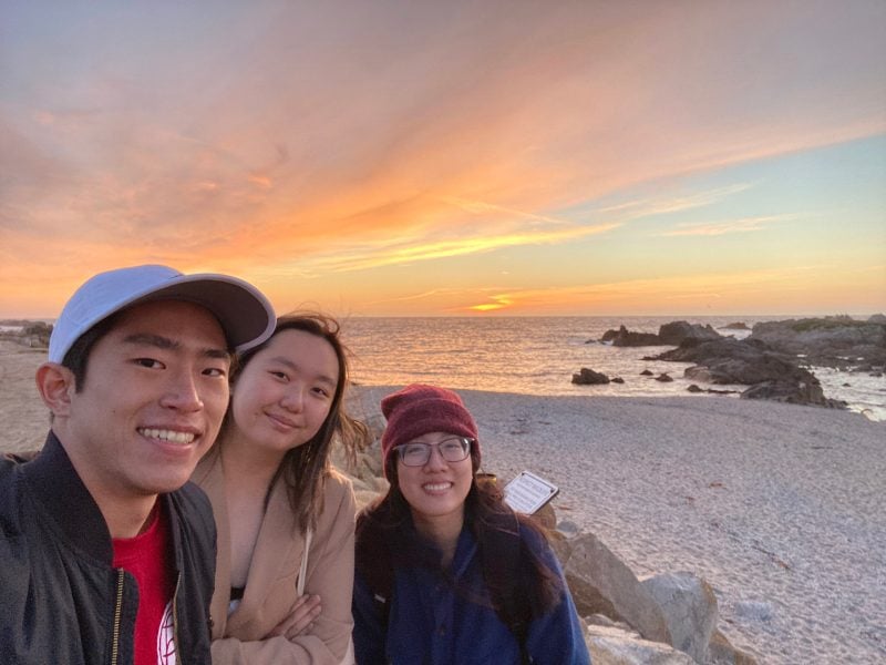 Three students smiling at a beach in front of a sunset.