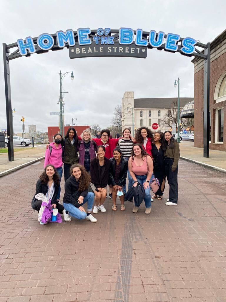 A group of students stand in the middle of a brick street under an arched sign which reads "Home of the Blues, Beale Street".