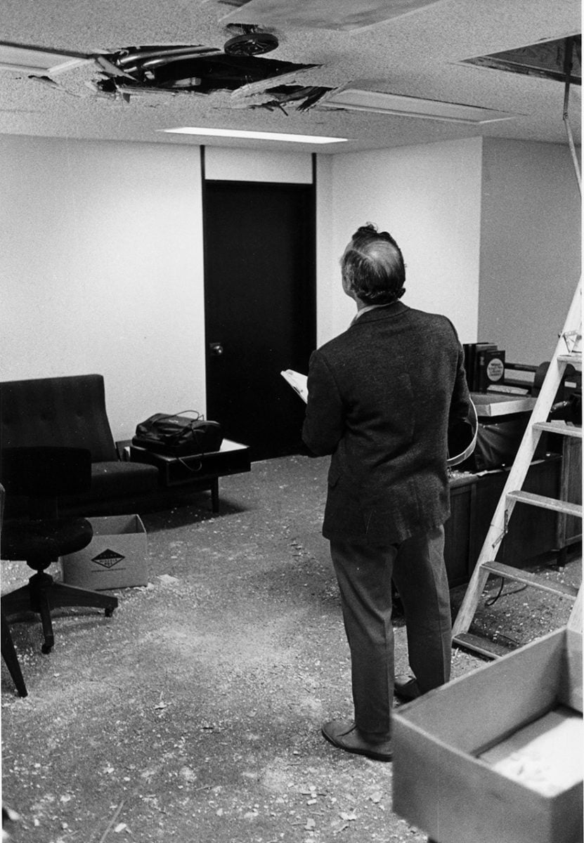 A balding man in a suit looks up at a hole in the ceiling.