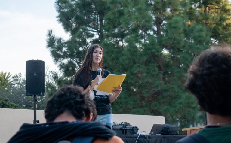 A woman holding papers and a microphone stands before a crowd, speaking to them