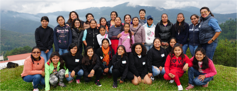 About thirty young girls and women pose for the camera, some kneeling and some standing behind. They stand on a green hill overlooking a valley in Tlahuitoltepec, Mixe, Oaxaca