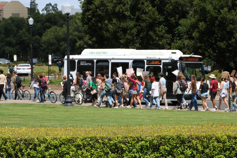 Protestors walk alongside the sidewalks of Jane Stanford Way. Many individuals hold signs, and many walk with their bikes. A shuttle bus is in the background. 