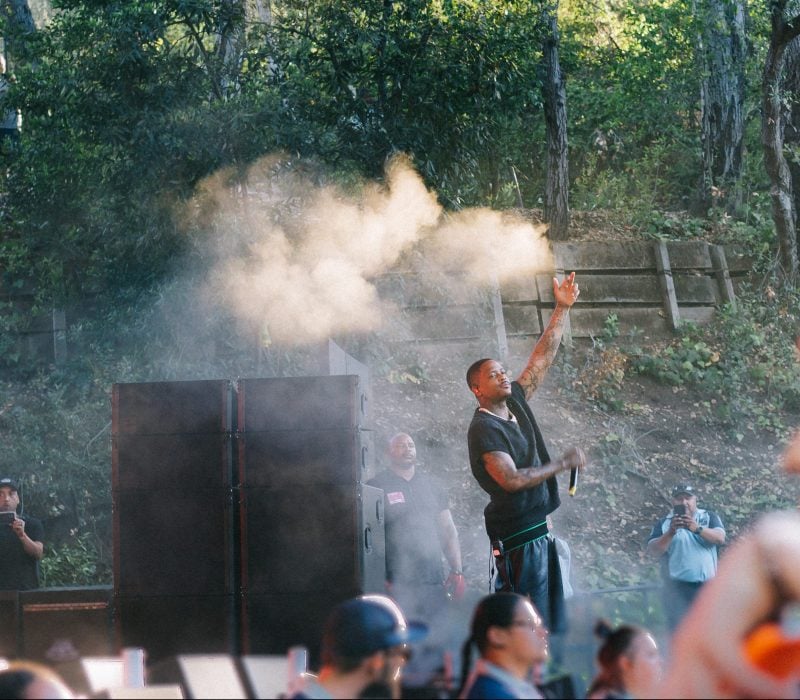 Blackfest headliner YG performs on stage. There is smoke in the air and he stands with his left hand up, looking at the crowd.