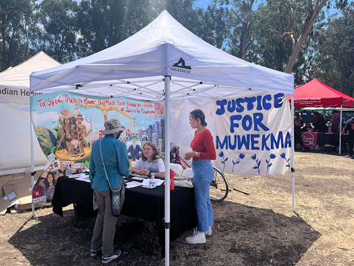 Students from Justice for Muwekma sit at a table under a tent, with a large sign that reads "Justice for Muwekma" behind them. They speak to a visitor. 