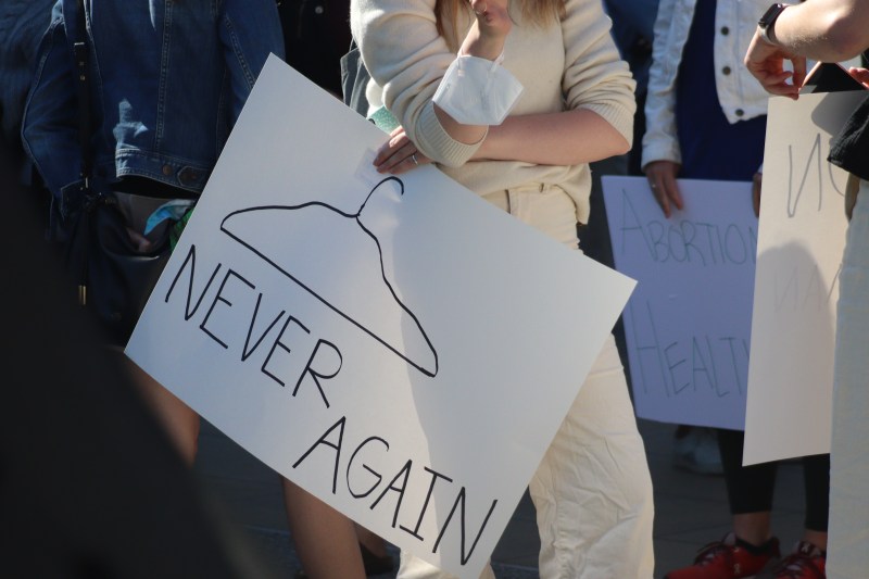 An attendee holds a white poster decorated with a drawing of a hanger and text reading "NEVER AGAIN" written in Black ink.