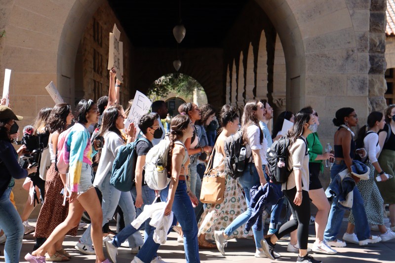 Protest attendees walk through the columns of main quad holding signs in support of reproductive rights. A videographer stands on the left side of the image, capturing the protest.