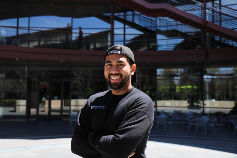 A man wearing a black shirt and black backwards baseball cap, standing in front of a glass building, with his arms crossed and smiling at the camera.