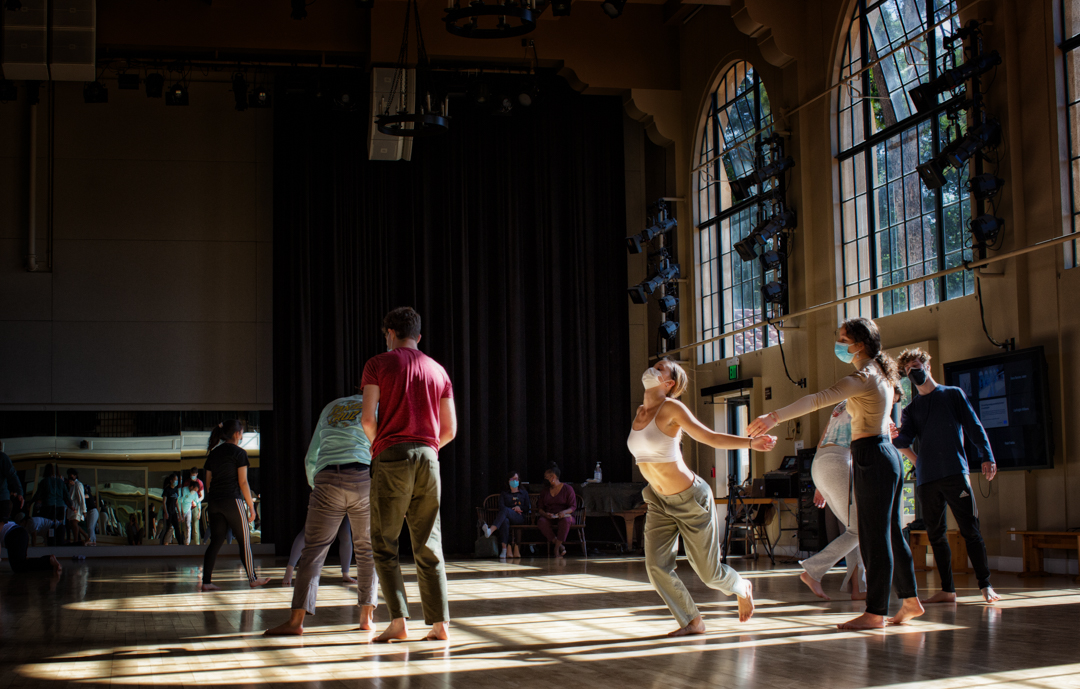 Groups  of students dance and interacting, the room they are in is spacious with really tall ceiling and arch windows. The person in the middle almost falls, anchored by their foot, with their dance partner reaching their arm out as if to try to catch them. Other students also pair up and interact.