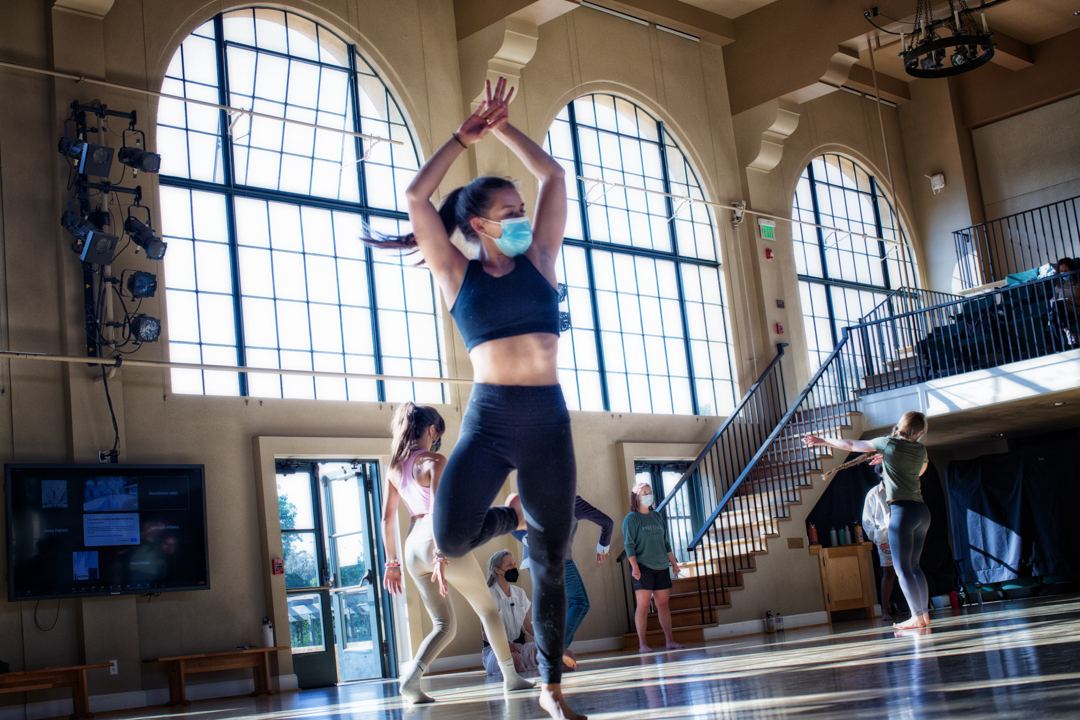 Person in black cropped top and black yoga pants stretched their arms up, one leg lifted, their ponytail swayed with their movement