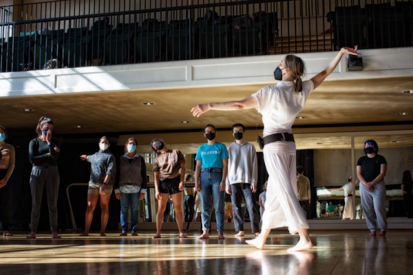 The person in white short-sleeved shirt and white flowy pants stretches her arm in an elegant arch, her right foot ahead of her left. The students stand behind her, admiring the elegant dance as they stood in an atrium with wooden floor and the shadow of the window casting down