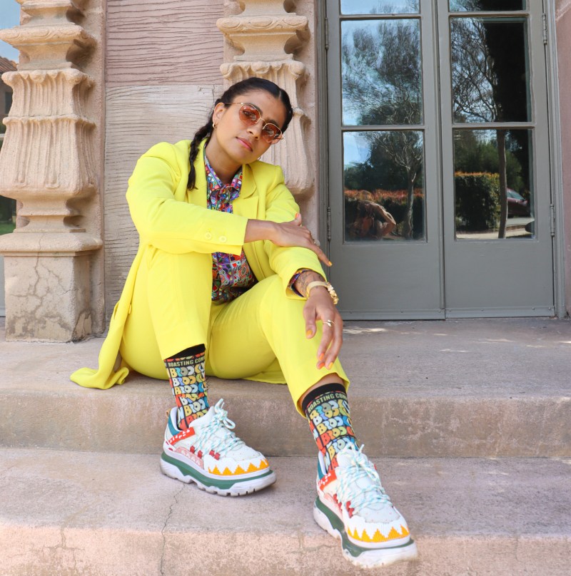 Madame Gandhi dressed in a yellow pantsuit and shade sits on the steps of the Center for Computer Research in Music and Acoustics