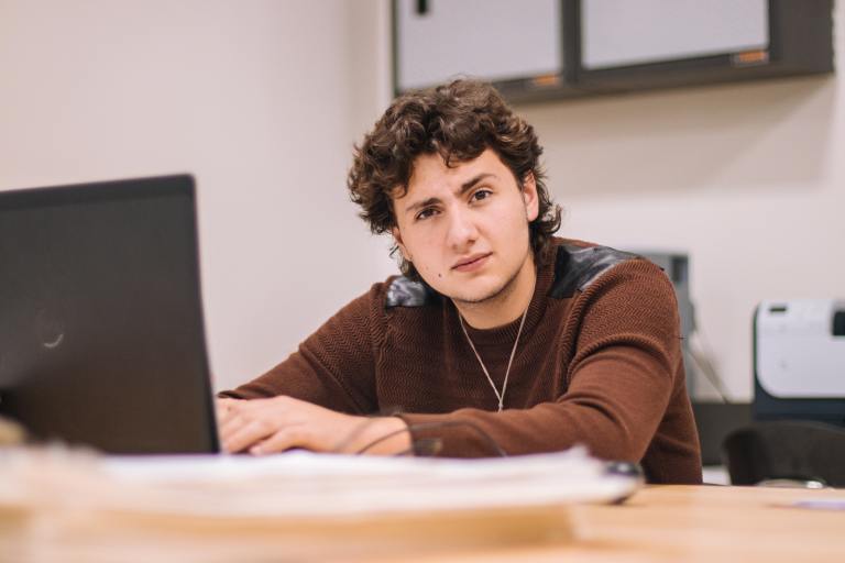 a sweatered person with long curly hair looks quizzically at the camera while typing at their laptop on a desk