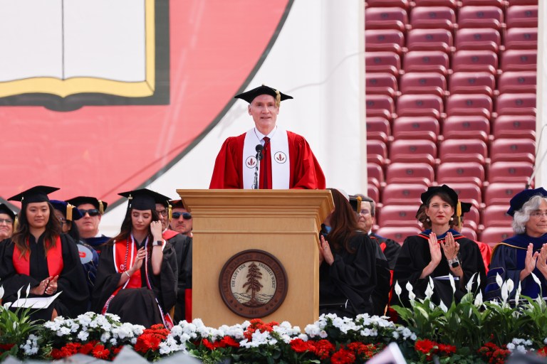 Marc Tessier-Lavigne addresses graduates from a podium, with faculty members in the back.