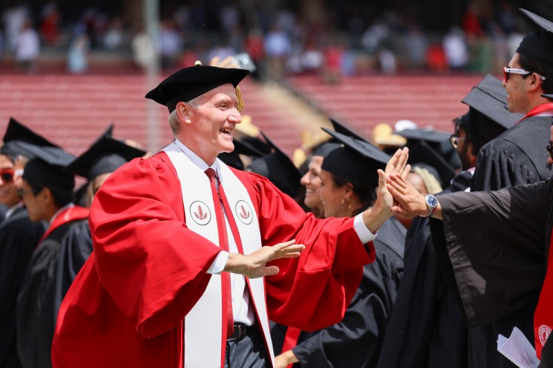 Stanford President Marc Tessier-Lavigne, wearing a red graduation gown and white stole, high-fives new grads