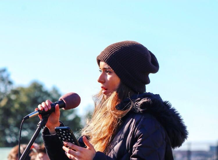 Photo of a Sophia Kianni in a dark brown beanie and black coat holds her phone in her hand and speaks into a microphone stand.