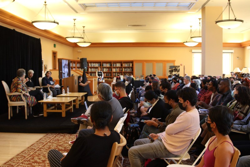 Panelists sit on the left side on a platform and rows of audience members sit on the right.