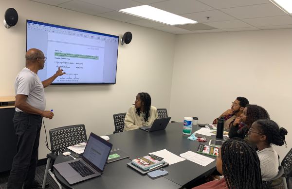 Biratu stands at the front of the room next to a screen lecturing; five students are watching.