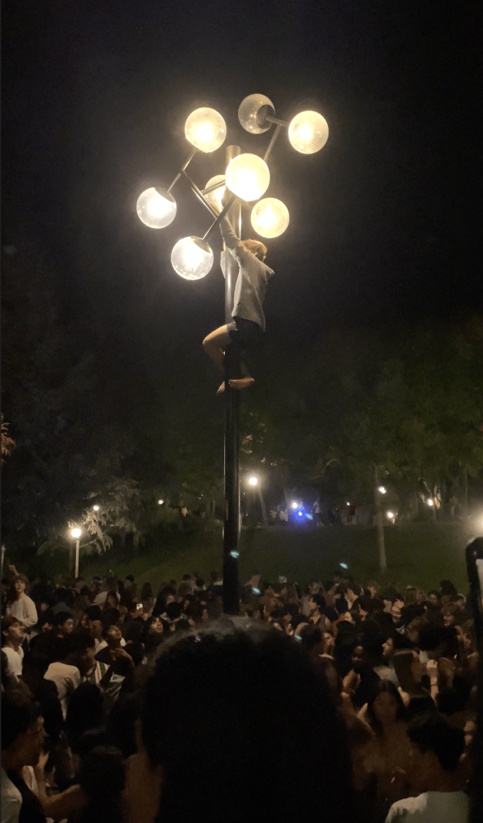 Students look on in Terman Fountain as one student in a grey sweatshirt climbs a light pole in Terman Fountain